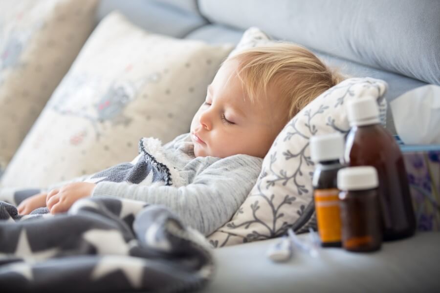 Przeziębiony chłopczyk leży w łóżku, obok stoją leki, chusteczki higieniczne i termometr.