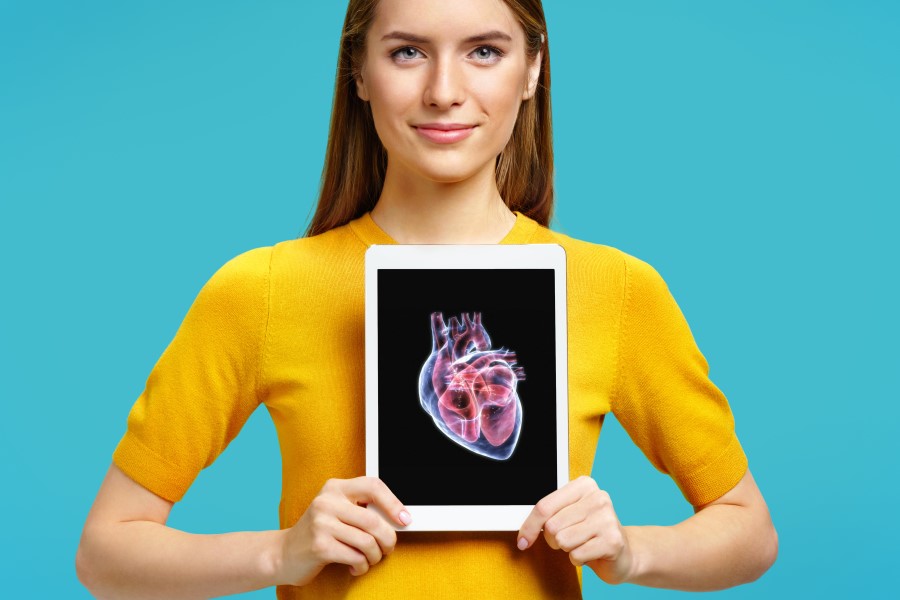 Młoda kobieta trzyma tablet na wysokości klatki piersiowej. Na ekranie wyświetla się model serca.