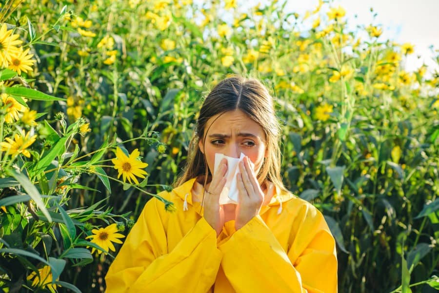 Kobieta w żółtej kurtce, pośród kwiatów. Wydmuchuje nos, bo cierpi na katar alergiczny.