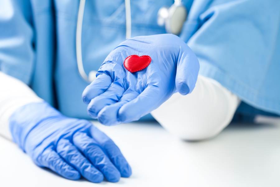 Lekarz ubrany na niebiesko, w niebieskich rękawiczkach. Trzyma na dłoni małe, czerwone serce.