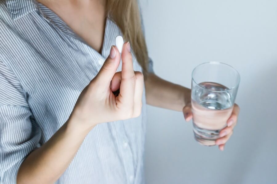 Kobieta w jednej dłoni trzyma szklankę, w drugiej tabletkę przeciwbólową.