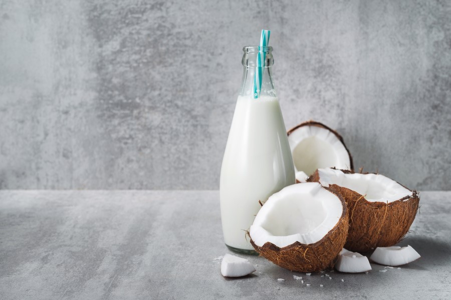 Mleko kokosowe w szklanej butelce oraz rozłupany orzech kokosowy na szarym tle.