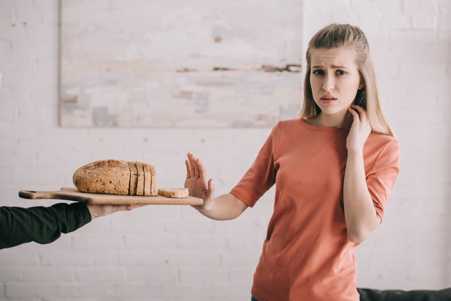 Dziewczyna z alergią pokarmową odsuwa od siebie deskę, na której leży pokrojony chleb.