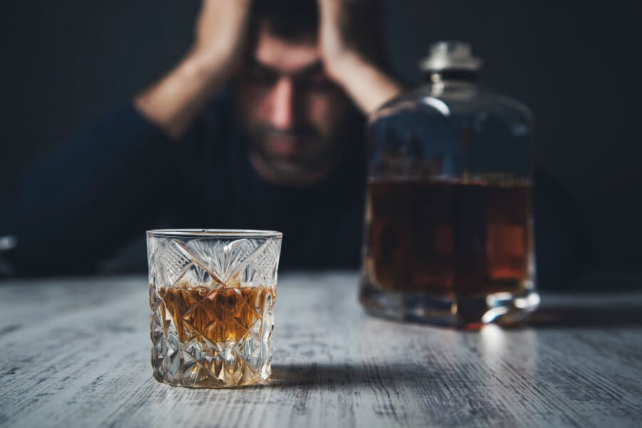 Mężczyzna dotknięty alkoholizmem siedzi przy stoliku, na którym stoi butelka i szklanka z alkoholem.