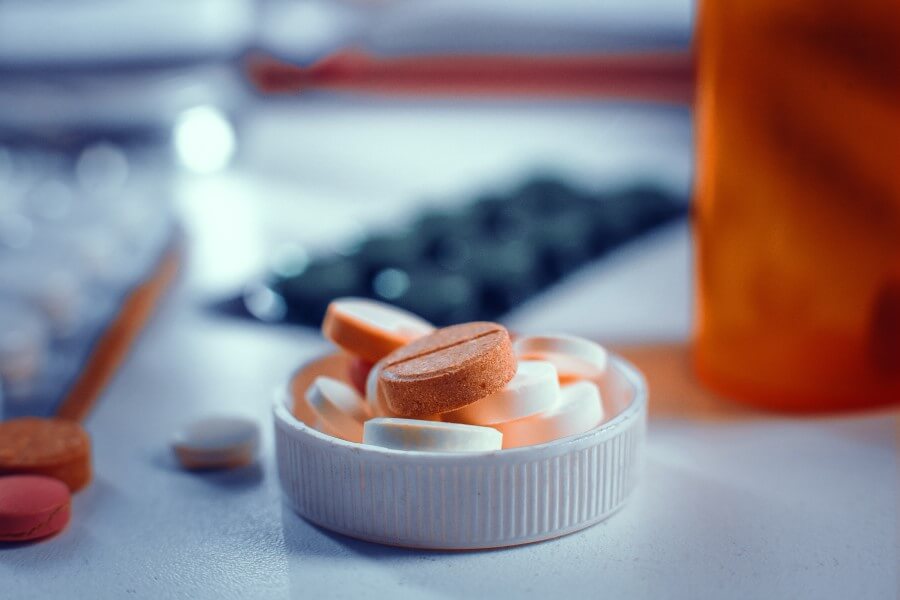 Białe i pomarańczowe tabletki w nakrętce po butelce leku, w tle blistry leków.
