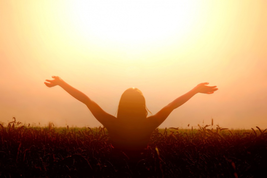 Kobieta stoi na polu pszenicy, na tle zachodzącego słońca, wyciąga ręce ku górze.