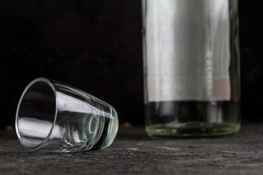 Przewrócony kieliszek leży na stole, w tle pusta butelka po alkoholu