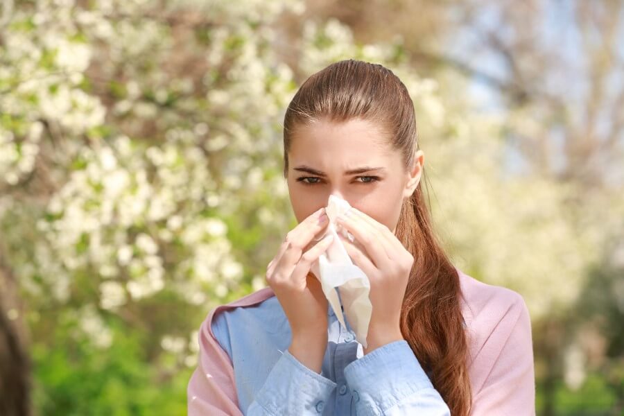 Kobieta stoi w kwitnącym sadzie i wydmuchuje nos, bo ma alergię na pyłki.