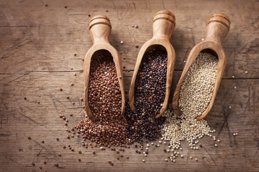 Trzy rodzaje quinoa (komosy ryżowej) w drewnianych łopatkach i częściowo rozsypane na blacie.