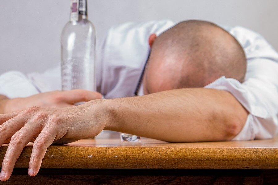Mężczyzna pod wypływem alkoholu, opiera głowę o blat stołu. Ręką trzyma butelkę z alkoholem.