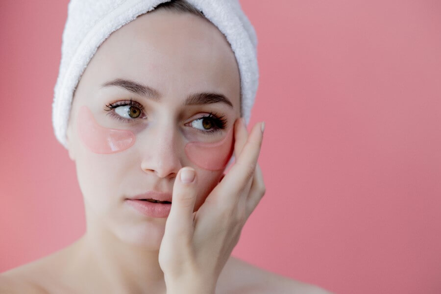 Kobieta po kąpieli, z ręcznikiem na głowie. Nakłada specjalne płatki niwelujące worki pod oczami.