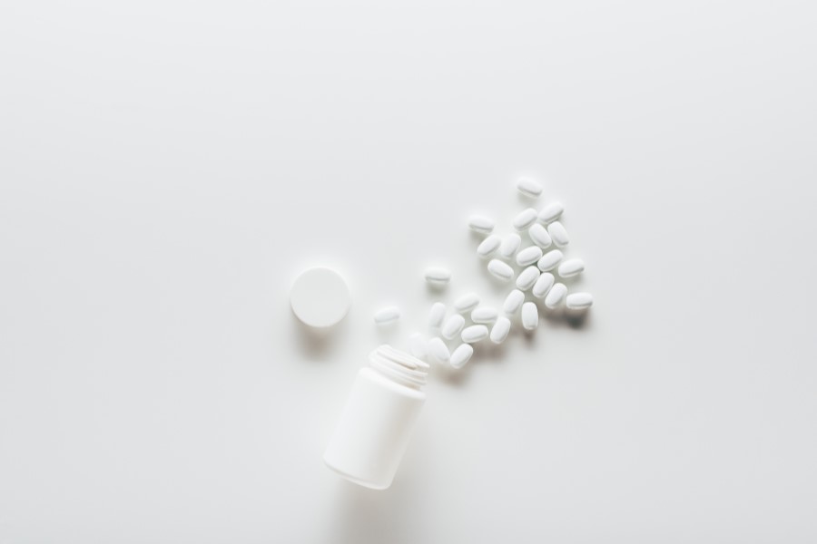 Biała butelka i wysypane z niej białe, owalne tabletki na białym tle.